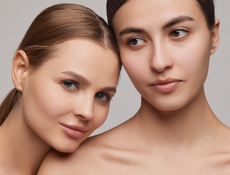 Zwei Frauen mit glatter Haut stehen eng beieinander vor einem grauen Hintergrund.